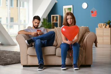 Sad young woman holding heart-shaped balloon near indifferent man playing with mobile phone at home. Relationship problems