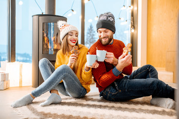 Young couple dressed in bright sweaters sitting together with hot drinks near the fireplace in the modern house in the mountains durnig winter time