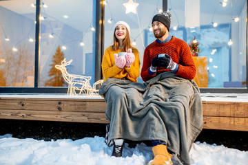 Young couple warming up with plaid and hot drinks sitting on the terrace of the modern house in the mountains durnig the winter holidays