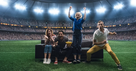 Group of students are watching a soccer moment, sitting on the couch located in the middle of the soccer stadium.