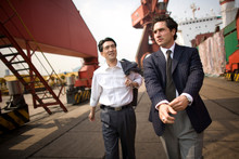 Young Adult Businessman Walking With A Mid-adult Male Colleague Along A Shipping Yard.