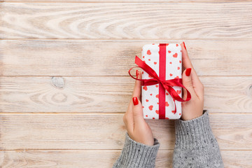 Woman hands give wrapped valentine or other holiday handmade present in paper with red ribbon. Present box, red heart decoration of gift on wooden table, top view with copy space for you design