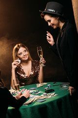 smiling attractive girls with glasses of champagne talking at poker table in casino