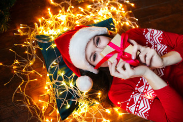 Close-up portrait of woman in Santa hat wrapped in christmas lights having fun and lying on floor