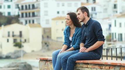 Poster - Serious couple of friends talking looking away sititng on a ledge on vacation in a coast town