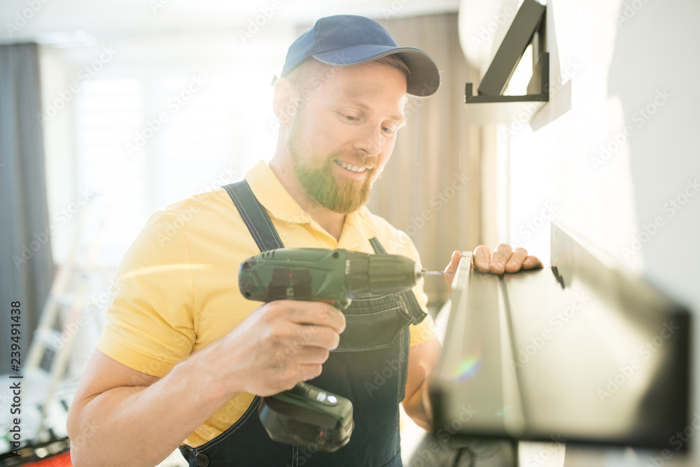 Cheerful optimistic handsome young bearded handyman in blue cap using cordless drill while screwing screw, he hanging shelf in apartment - obrazy, fototapety, plakaty 