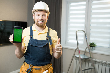 Smiling handsome young bearded guy with tool belt wearing white hardhat and denim overall holding smartphone and showing thumb-up while looking at camera and standing in modern flat