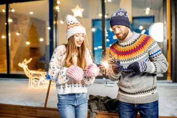 Young happy couple dressed in sweaters celebrating winter holidays with bengal fire in front of a beautiful decorated house