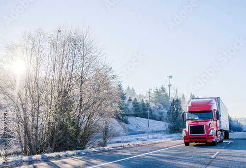 Bright red big rig semi truck with refrigerator semi trailer transporting cargo on straight winter highway frosty hill trees