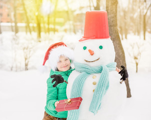 Wall Mural - portrait of smiling boy in red christmas hat holds gift box and hugs snowman. Empty space for text