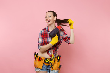 Wall Mural - Young beautiful fun handyman woman in plaid shirt, denim shorts, kit tools belt full of variety instruments holding power heat gun isolated on pink background. Female in male work. Renovation concept