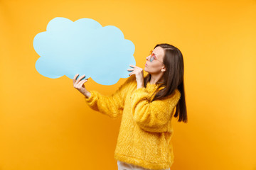 Brunette young woman in heart eyeglasses looking on empty blank blue Say cloud speech bubble in hands isolated on bright yellow background. People sincere emotions lifestyle concept. Advertising area.