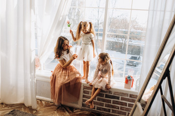 Wall Mural - Two little sisters dressed in the beautiful dresses and there young mother sit on the windowsill next to the mirror the winter is outside