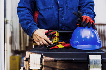 Wall Mural - Electrian engineer holding multimeter and tools in hand, standing behind the heavy duty tool box, image including power cord, Blue hard hat (helmet) and gloves.