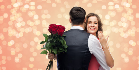 Wall Mural - love, couple, proposal and people concept - happy woman with engagement ring and bunch of roses hugging man over living coral background and festive lights