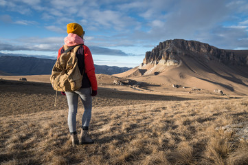 Wall Mural - Portrait from the back of a girl traveler in a jacket with a cap and a backpack stands on the background of an epic landscape with rocks