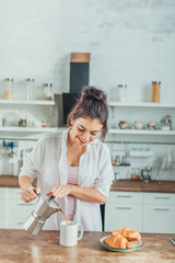 smiling pretty girl pouring coffee into cup at wooden table with croissants in kitchen at home