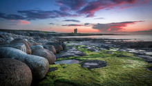 Big Round Rocks And Stones With Green Seaweed At The Coast Of Easkey Beach, Ireland, With In The Background The Ruin Of A Castle-tower Under The Blue And Purple Sky After Sunset