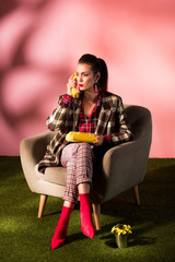 Wall Mural - fashionable girl in checkered suit posing in armchair with rotary phone on pink background