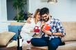© Mediteraneo - Man surprising his girlfriend with a gift on the couch at home in the living room