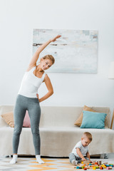 smiling woman stretching, smiling and looking at camera and adorable toddler boy playing with multicolored cubes on carpet at home