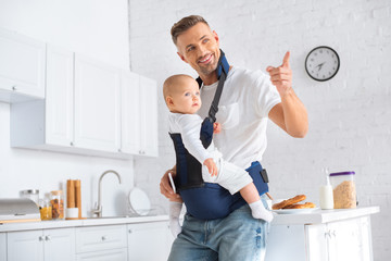 Wall Mural - cheerful father holding infant daughter in baby carrier and pointing with finger