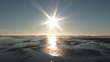 Wall Mural - Sea water carves through sandy beach with sun centered in clear blue sky at the Pacific Ocean.
