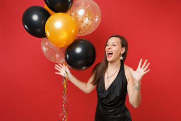 Joyful young girl in little black dress celebrating, spreading hands, holding air balloons isolated on red background. International Women's Day, Happy New Year, birthday mockup holiday party concept.