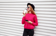 © guas - Stylish young smiling woman drinks coffee holds phone in pink knitted sweater on white wall background