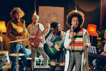 Canvas Print - Mixed race woman singing. In background band playing instruments. Home studio interior.