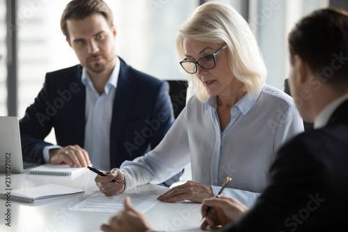 Fotografía Businessmen sitting at desk headed by middle aged serious concentrated female in eyeglasses checking agreement document before signing it