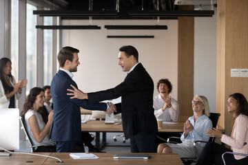 Canvas Print - Mature director company owner congratulating young specialist handshaking standing at coworking area, diverse multiracial coworkers different colleagues applauding rejoicing for success of colleague