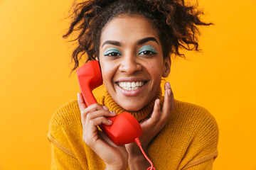 Wall Mural - Portrait of positive african american woman with afro hairstyle smiling and holding red handset, isolated over yellow background