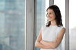 © fizkes - Successful millennial smiling brunette businesswoman standing in contemporary office with hands crossed on chest, looking through window at big city modern buildings, thinking planning new projects