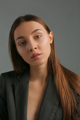 Portrait of pretty young woman of natural beauty, without makeup, full lips, long straight brown hair with parting, looking at camera, wearing classic jacket, isolated on grey background