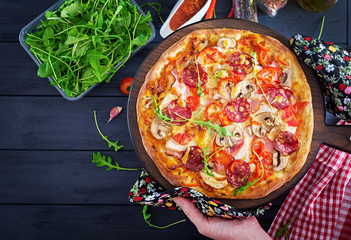 Wall Mural - Fresh Italian pizza with chicken fillet, mushrooms, ham, salami, tomatoes, cheese on a black background in the hands. Italian food.