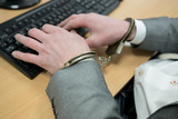 Fototapeta Zachód słońca - Detail of business person hand tied with handcuffs to workplace, keyboard and monitor in background.
