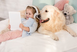 Fototapeta Przestrzenne - adorable child listening music with tablet and leaning on cute golden retriever on bed in children room