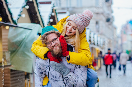 Fotografía Couple in warm bright outwear, man giving piggyback to woman in street, outdoor