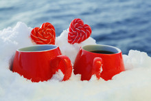 Two Red Mugs With Hot Tea Or Coffee And Lollipops Hearts In The Snow. Picnic On A Frosty Sunny Day On A River Or Sea Background On Valentine's Day. The Concept Of Celebrating Valentine's Day