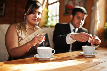 Wall Mural - Elegant and fashionable indian friends couple of woman in saree and man in suit sitting on cafe and drinking cappuccino.