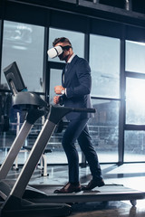 Wall Mural - side view of businessman in suit and virtual reality headset exercising on treadmill in gym