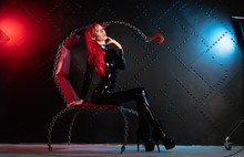 Dangerous Cruel Fetish Girl With Red Hair Posing In Black Bdsm Latex Clothes At The Chair From The Chains In The Style Of Sadomasochism