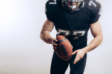 Wall Mural - Athletic american football player in uniform and headgear holding oval ball posing isolated over white background.
