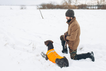 Wall Mural - young ambitious man and his dog sitting on the snow . side view shot. hunter resting with a pet outdoors