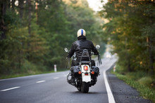Back View Of Biker In Black Leather Jacket And White Helmet Riding Motorbike Along Hilly Road Between Tall Green Trees. Active Lifestyle, Love To Adventures Concept.