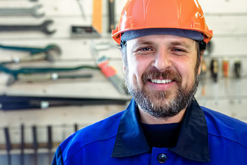 A man with a beard in a helmet and work clothes is smiling a snow-white smile against the background of a stand with tools. Portrait of a worker in workwear with copy space.