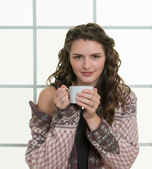 Pretty woman in window wearing cozy sweater drinking hot beverage mug