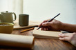 © DragonImages - Close-up of female hands holding pencil and writing daily routine in her notebook at the table with cup of tea
