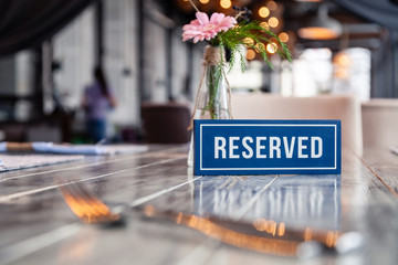 Closeup wooden blue white rectangular plate with word Reserved standing on gray vintage table in restaurant near to setting, napkins, fork, knife, vase with flowers, glasses, ceramic white plates